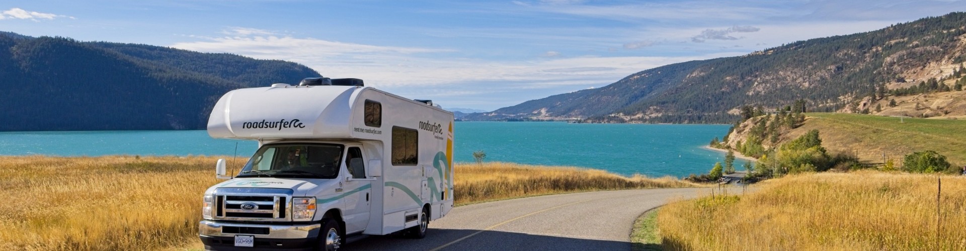 Class C RV driving along a winding lakeside road with blue water, fields, and mountains in the background, illustrating RV rentals near me