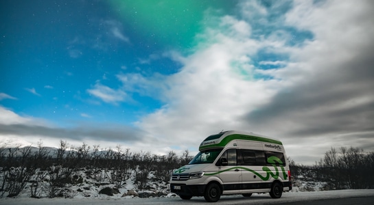 Big green and white RV standing on a road in Sweden with the northern lights visible in the sky