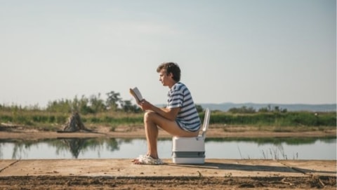 Person sitting on a portable RV toilet outdoors near a lake during camping
