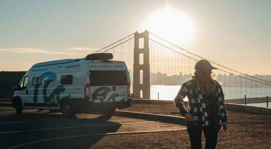 Woman standing near a parked RV rental with the Golden Gate Bridge in the background at sunrise in San Francisco.