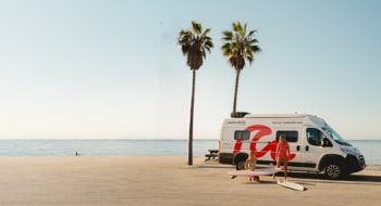 scenic beachside setting with a camper van parked near palm trees, two people in swimwear handling surfboards, and the ocean stretching into the horizon under a clear sky