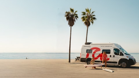 scenic beachside setting with a camper van parked near palm trees, two people in swimwear handling surfboards, and the ocean stretching into the horizon under a clear sky
