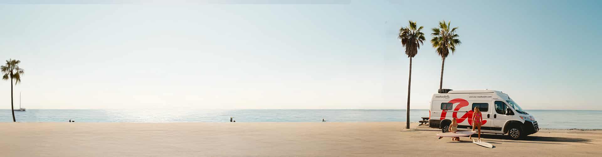 scenic beachside setting with a camper van parked near palm trees, two people in swimwear handling surfboards, and the ocean stretching into the horizon under a clear sky