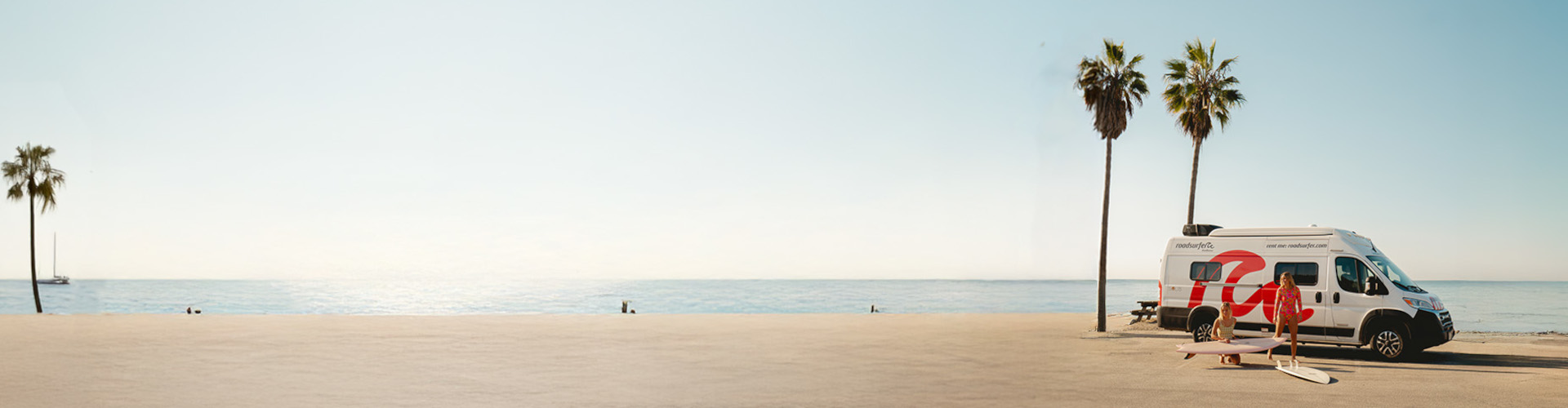 scenic beachside setting with a camper van parked near palm trees, two people in swimwear handling surfboards, and the ocean stretching into the horizon under a clear sky