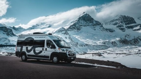 A small RV rental parked along a snowy mountain pass under a bright blue sky.