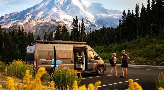 A small RV rental parked at a scenic mountain viewpoint with two hikers getting ready for their trip.