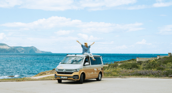 A young woman sits on her gold and white campervan parked by the Costa Brava sea, arms outstretched and cheering.