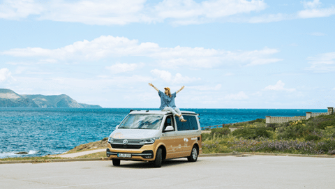 A young woman sits on her gold and white campervan parked by the Costa Brava sea, arms outstretched and cheering.