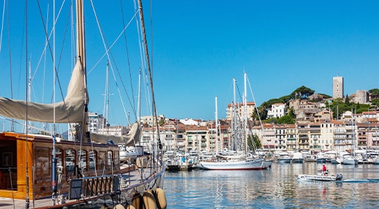 In the foreground you can spot sailing boats, one brown one in the left corner and white ones in the back. Behind all of the boats you can see the city Cannes and a hill in the background.