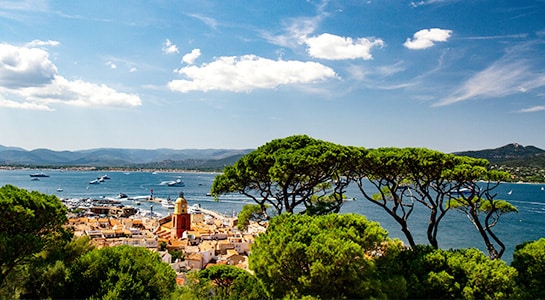 View over the city of Saint Tropez. You can see the blue sky with a few white clouds in the sky. In the foreground you can spot green trees blocking and through those trees you can see parts of the city and the sea and boats.