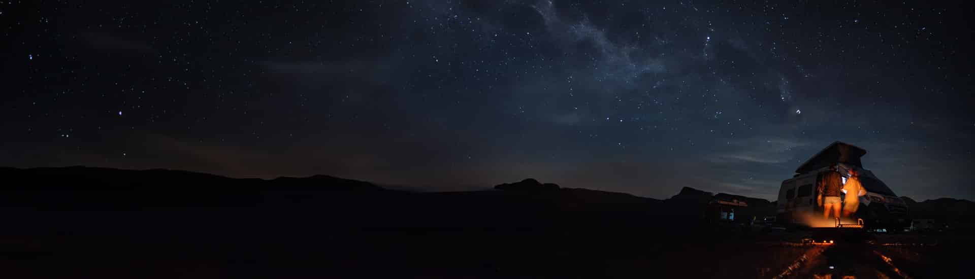 A star-filled night sky stretches over silhouetted mountains as two people stand beside a lit camper van near a small campfire.