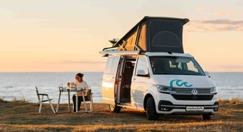 A compact camper van parked by the seaside at sunset, with its pop-up roof extended and side door open. A person sits at a small outdoor table beside the van, enjoying a quiet moment with a drink while overlooking the ocean. The scene conveys freedom, travel, and a minimalist lifestyle surrounded by nature, with warm golden light illuminating the coastal landscape.