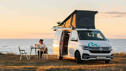 A compact camper van parked by the seaside at sunset, with its pop-up roof extended and side door open. A person sits at a small outdoor table beside the van, enjoying a quiet moment with a drink while overlooking the ocean. The scene conveys freedom, travel, and a minimalist lifestyle surrounded by nature, with warm golden light illuminating the coastal landscape.