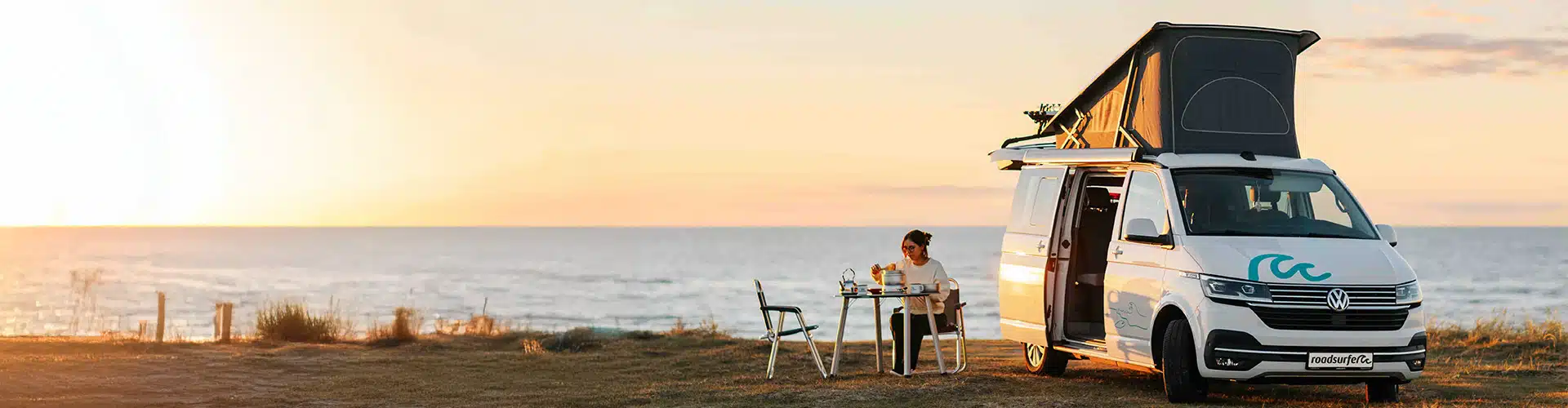 A compact camper van parked by the seaside at sunset, with its pop-up roof extended and side door open. A person sits at a small outdoor table beside the van, enjoying a quiet moment with a drink while overlooking the ocean. The scene conveys freedom, travel, and a minimalist lifestyle surrounded by nature, with warm golden light illuminating the coastal landscape.