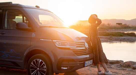A woman leaned on a campervan near to a lake looking into the sunset