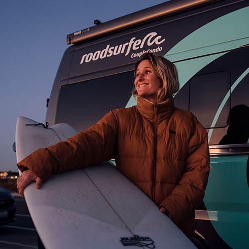 Girl standing in front of a roadsurfer RV, holding a surfboard and smiling