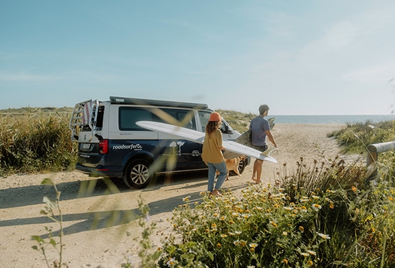 Two surfers leaving their camper on the beach and walking towards the sea with their surfboards.