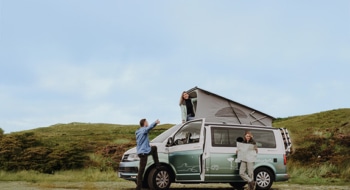 Two people standing in front of a campervan, while one is holding a card. Another woman is looking out of the open rooftop tent.