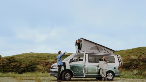 Two people standing in front of a campervan, while one is holding a card. Another woman is looking out of the open rooftop tent.