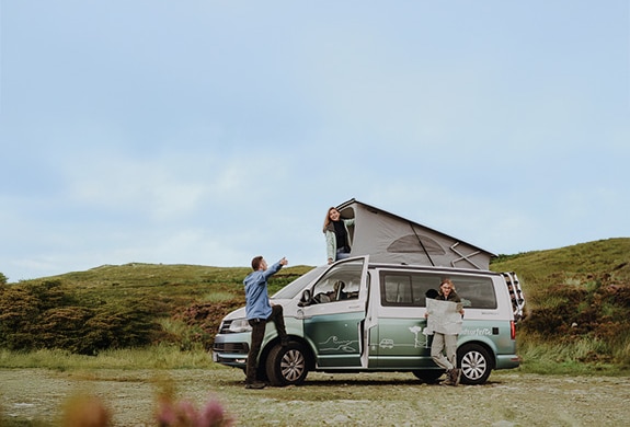 Two people standing in front of a campervan, while one is holding a card. Another woman is looking out of the open rooftop tent.