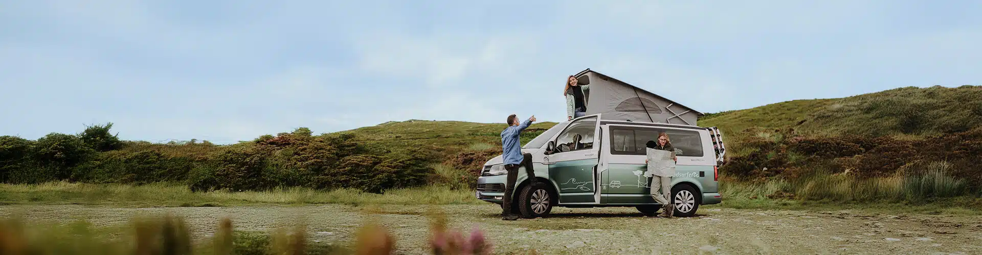 Two people standing in front of a campervan, while one is holding a card. Another woman is looking out of the open rooftop tent. 