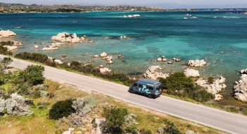 A big size campervan driving on a road next to a sea with turquoise water.