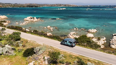 A big size campervan driving on a road next to a sea with turquoise water.