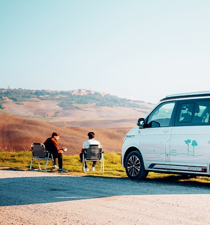Two friends sitting on chairs in front of their campervan enjoying coffee
