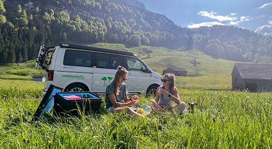 Two girls sitting in a field in front of a roadsurfer campervan on a sunny day