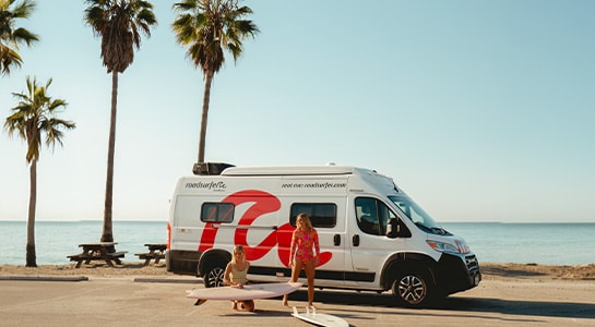 Two girls preparing their surfboards in front of their parked boxvan at the beach.