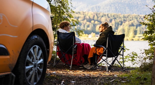 Couple sitting on camping chairs, each covered by a blanket, in front of a lake right next to a parked orange campervan