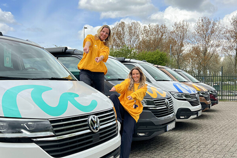 Two women pointing excitedly at roadsurfer campervans parked in a row behind and in front of them