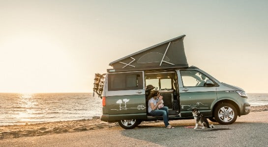 Mother and baby sit in the VW camper with their dog and watch the sunset