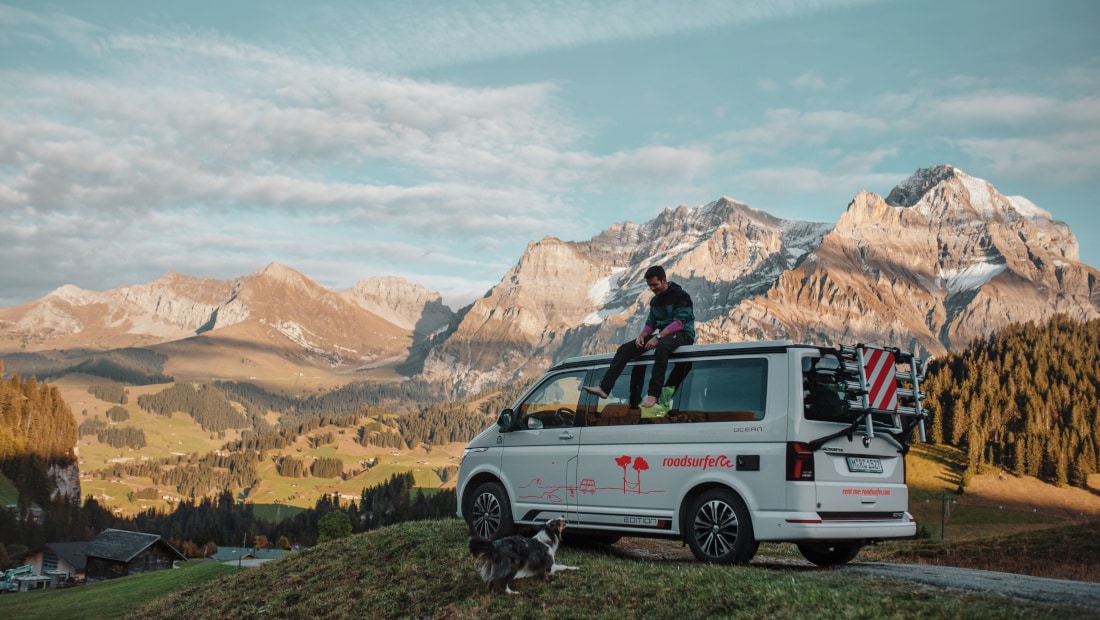 man sitting on a white van looking at his dog on the ground, mountains in the background
