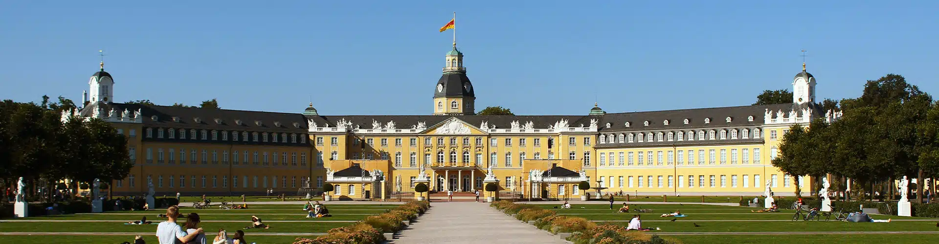 View of the castle in Karlsruhe on a sunny day with people either walking towards the castle or sitting on grass patches right in front of it.