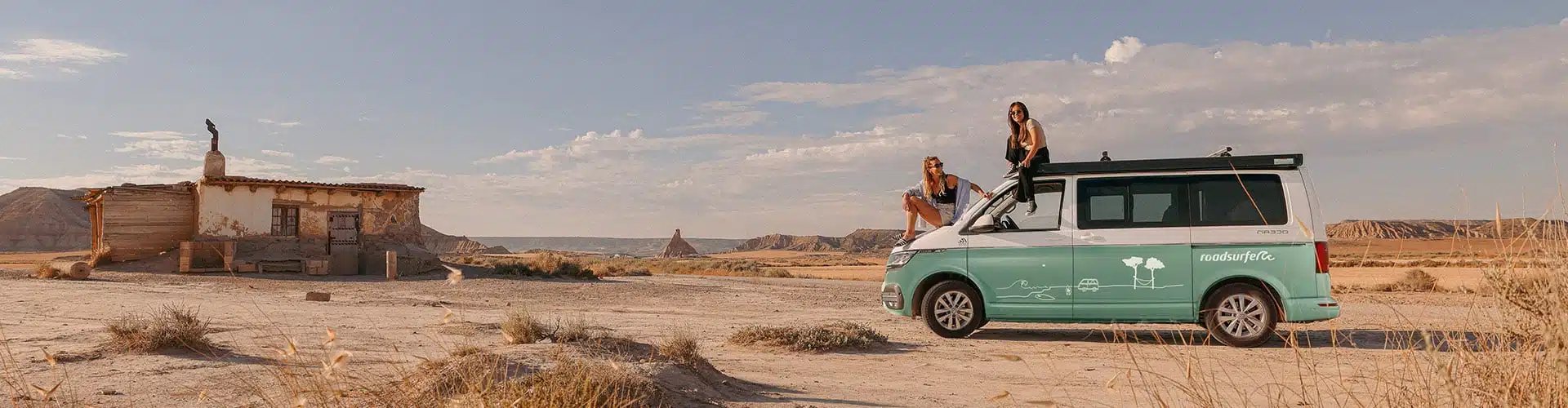 VW California camper van from roadsurfer in the desert sun, two smiling girls sitting on the hood and roof of the van