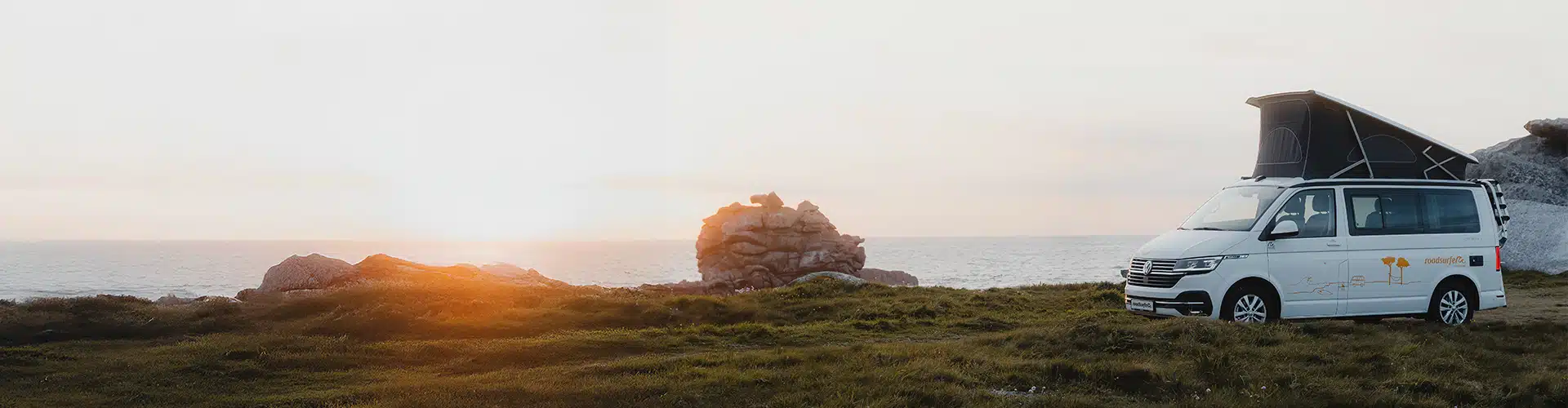 VW California Ocean campervan parked on a grassy coastal area at sunset, van overlooking the ocean with a pile of rocks nearby and the horizon bathed in warm sunset light