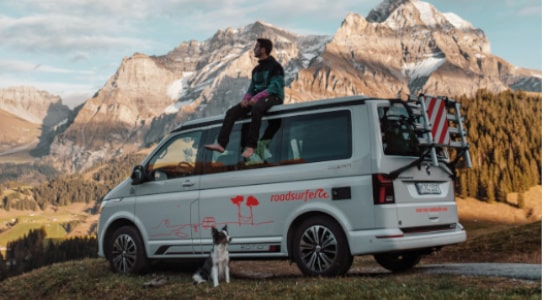 Man sits on the roof  of a VW California Ocean and looks a the view of the mountains whilst his dog waits on the ground