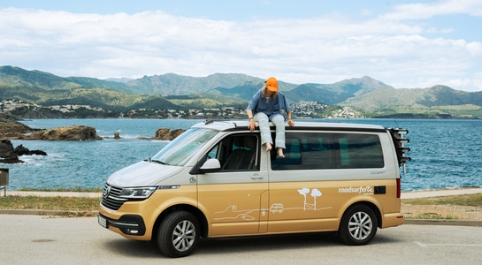 Young girl with a orange beenie is sitting on roof of a VW California camper standing at a parking next to the sea at the Costa Brava in Spain