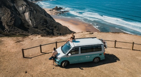 Camper van parked on a hill overlooking the ocean with a person sitting on the roof and another leaning against the front of the van while they take in the views of the Algarve coast