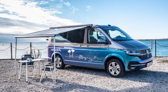Blue and grey VW campervan with open awning and camping table and chair set up in front of sea