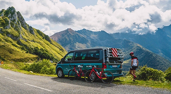 VW campervan standing on a road on the Alps with 2 bikes leaned on it, ready for a bike tour
