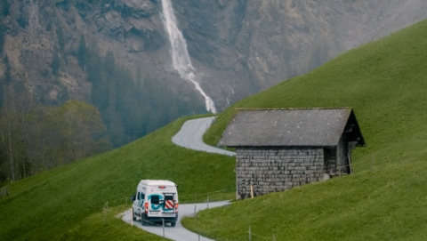 roadsurfer campervan driving on a road in Switzerland with a waterfall in the backgrounds comming out from the mountain