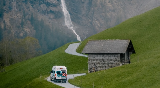 roadsurfer campervan driving on a road in Switzerland with a waterfall in the backgrounds comming out from the mountain