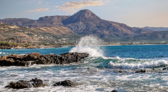 Waves crash dramatically against rugged coastal rocks with a mountainous backdrop on a sunny day in Crete.