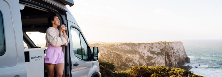 Woman standing in doorway of boxvan drinking a coffee and overlooking the view over the cliffs and the sea.