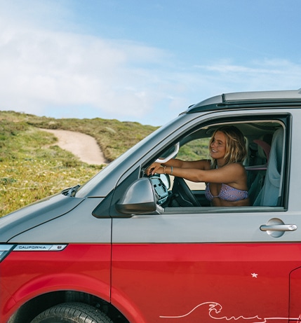 smiling woman in a bikini sitting in the driver's seat of a red and gray VW California camper van, parked in a scenic, grassy coastal area under a blue sky