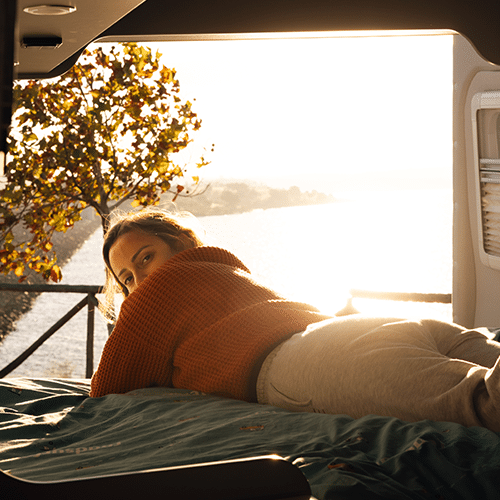 A young woman gazes back while reclining on the rear bed of a campervan, the open doors framing a breathtaking sunset