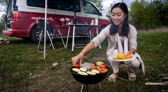 Woman cooking vegetables on a small barbecue and the campsite and a campervan visible in the background.