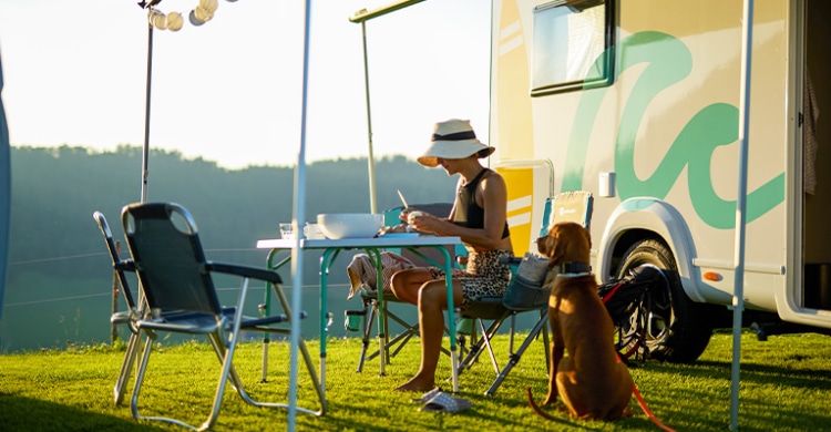 Woman cutting food on a camping table in front of her parked camper, with her dog sitting and watching her.